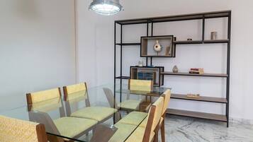 A dining area with a glass table and chairs, a metal-framed bookshelf, and a pendant light.