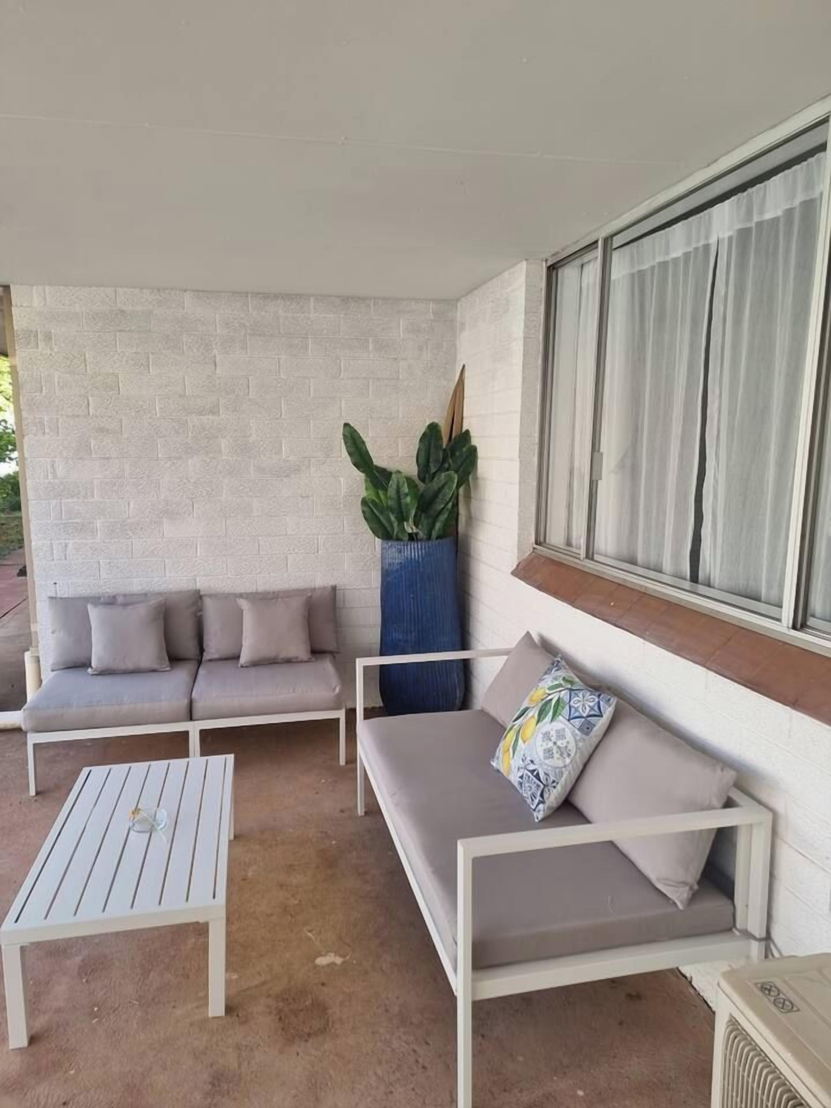 A covered outdoor seating area with a white brick wall, a large potted plant, and a window with curtains.