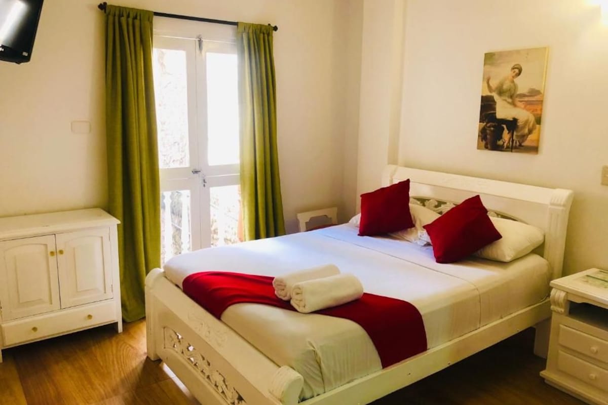 A hotel room with a bed, a nightstand, a television, and a painting on the wall.