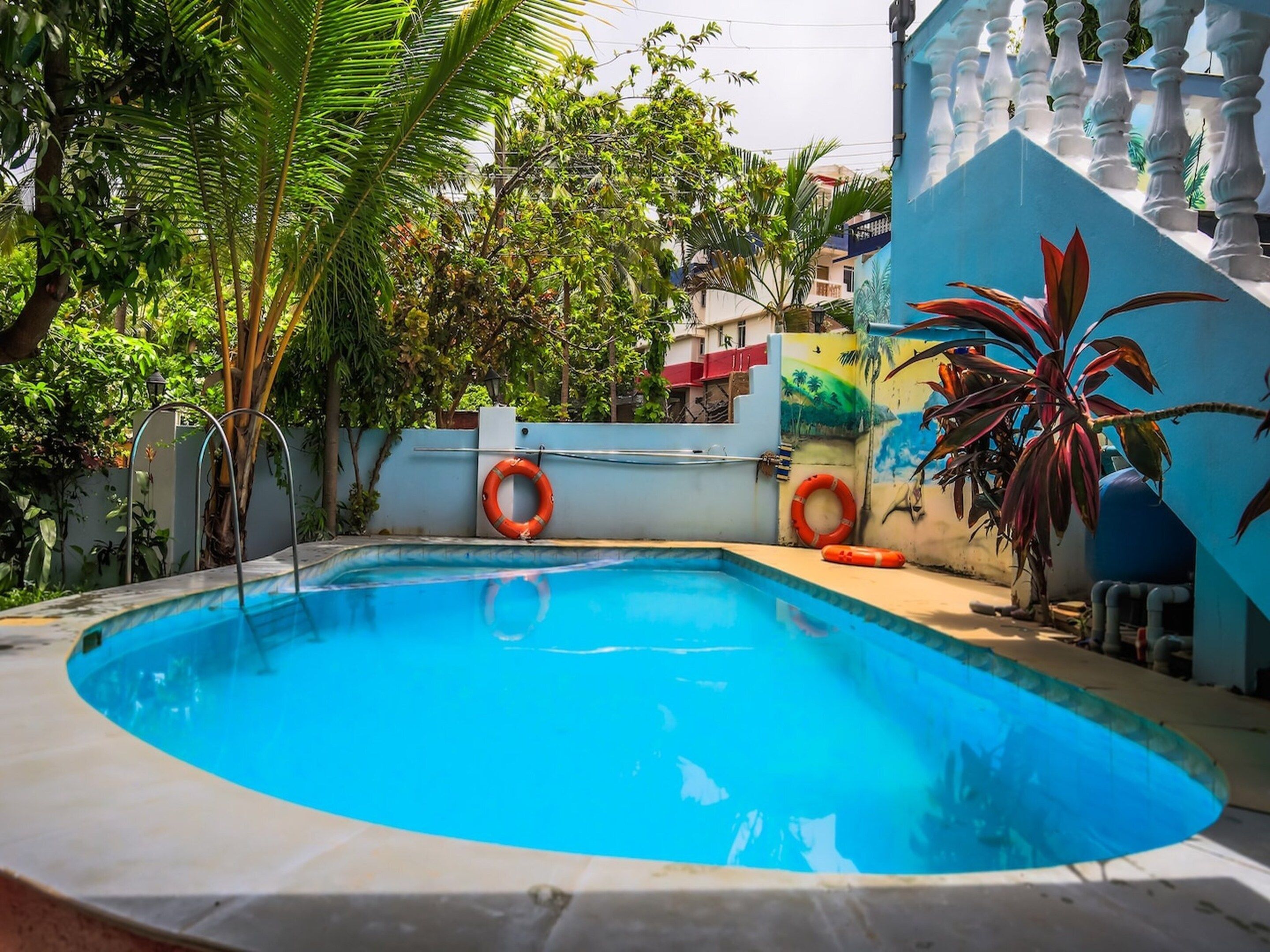 A swimming pool with a curved edge, surrounded by a white wall with a mural, and two orange lifebuoys attached to the wall.