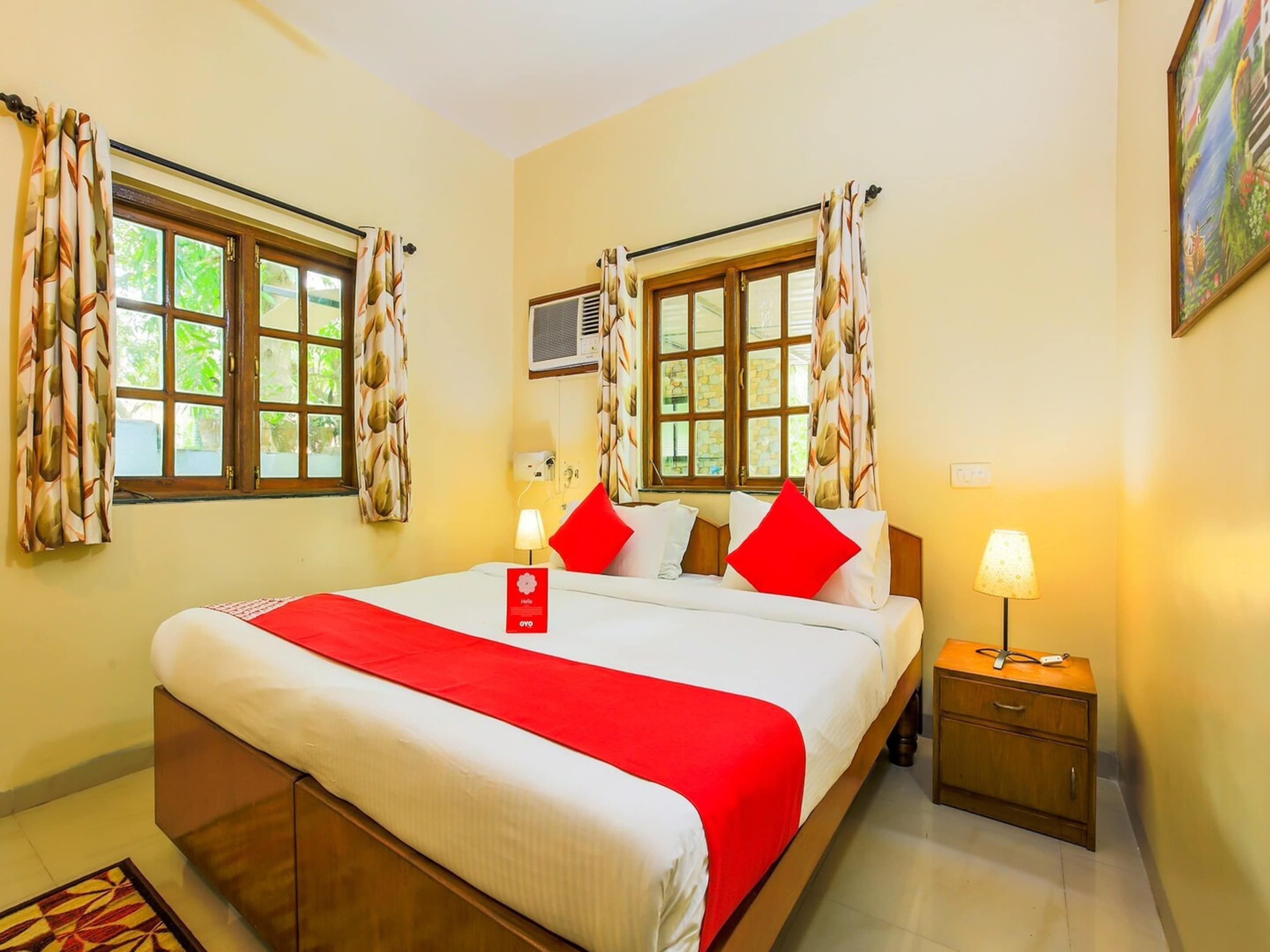 A hotel room with a bed, two bedside tables, a nightstand, a window with curtains, an air conditioning unit, and a painting on the wall.