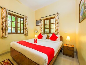 A hotel room with a bed, two bedside tables, a nightstand, a window with curtains, an air conditioning unit, and a painting on the wall. - Classic Tanisha Guest House (Marmagao)