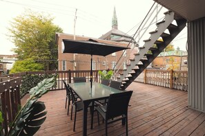 Eine Holzterrasse mit Tisch und Stühlen, eine Treppe und im Hintergrund ein Gebäude mit grüner Dachbegrünung.