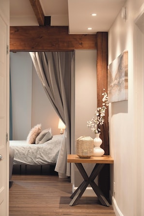 Une chambre à coucher avec un lit, une table de chevet ornée d’un vase et d’un panier, un plancher en bois et un plafond avec des poutres apparentes.