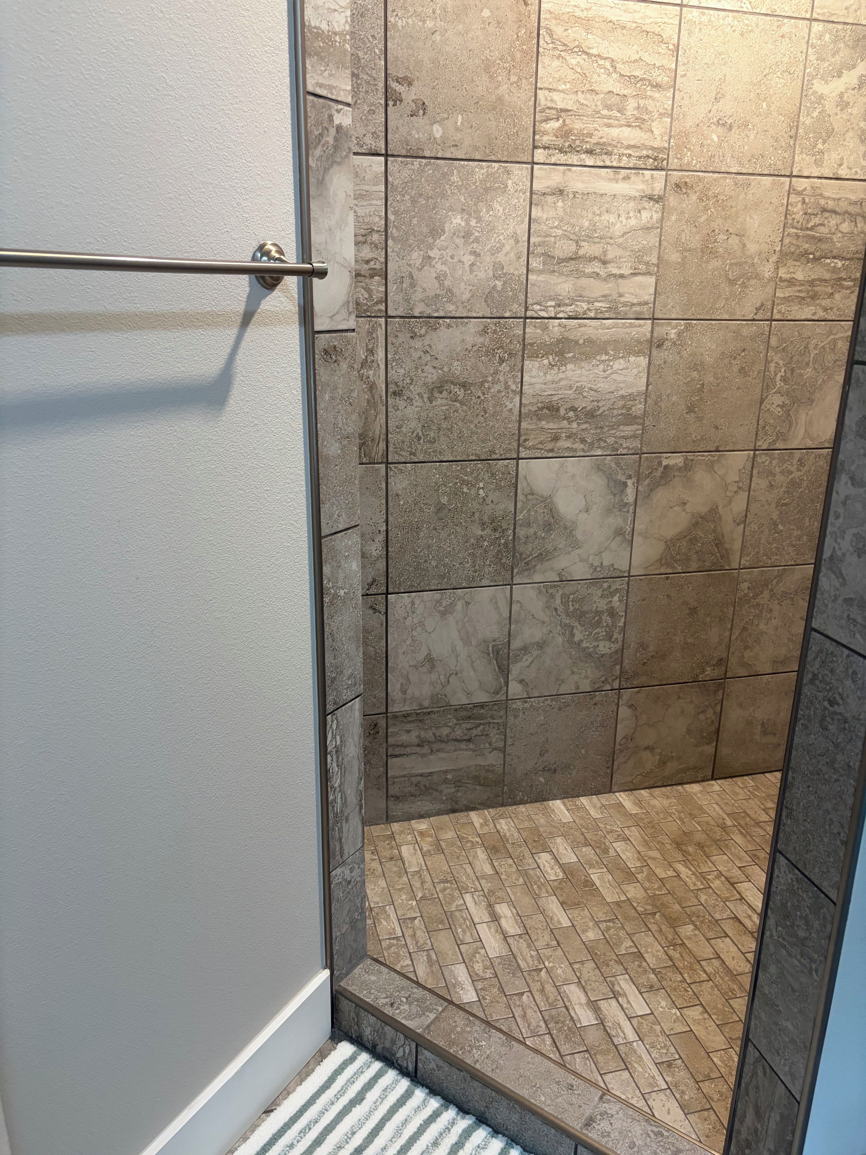 Un espace douche avec un mur et un sol carrelés, un porte-serviettes en métal et un tapis à rayures.