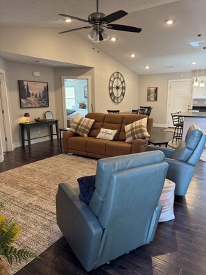 Un salon moderne avec un canapé marron, des fauteuils inclinables bleus, un grand tapis et un ventilateur de plafond.