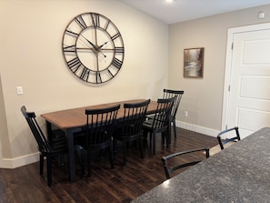 Un coin repas avec une table en bois et des chaises noires, une grande horloge murale et un tableau encadré accroché au mur.