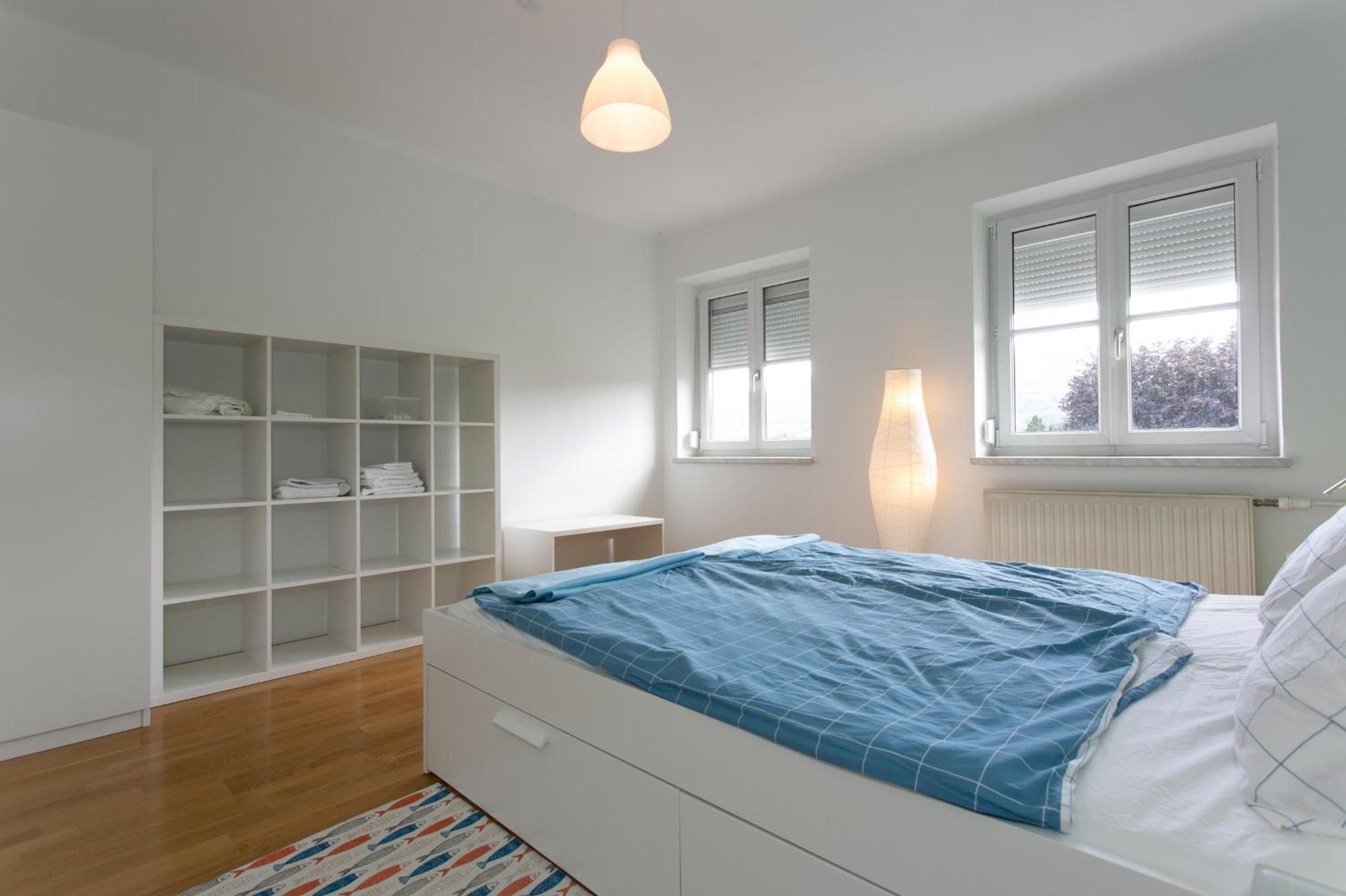 A bedroom with a bed, a white wardrobe, a window with blinds, and a hanging light fixture.