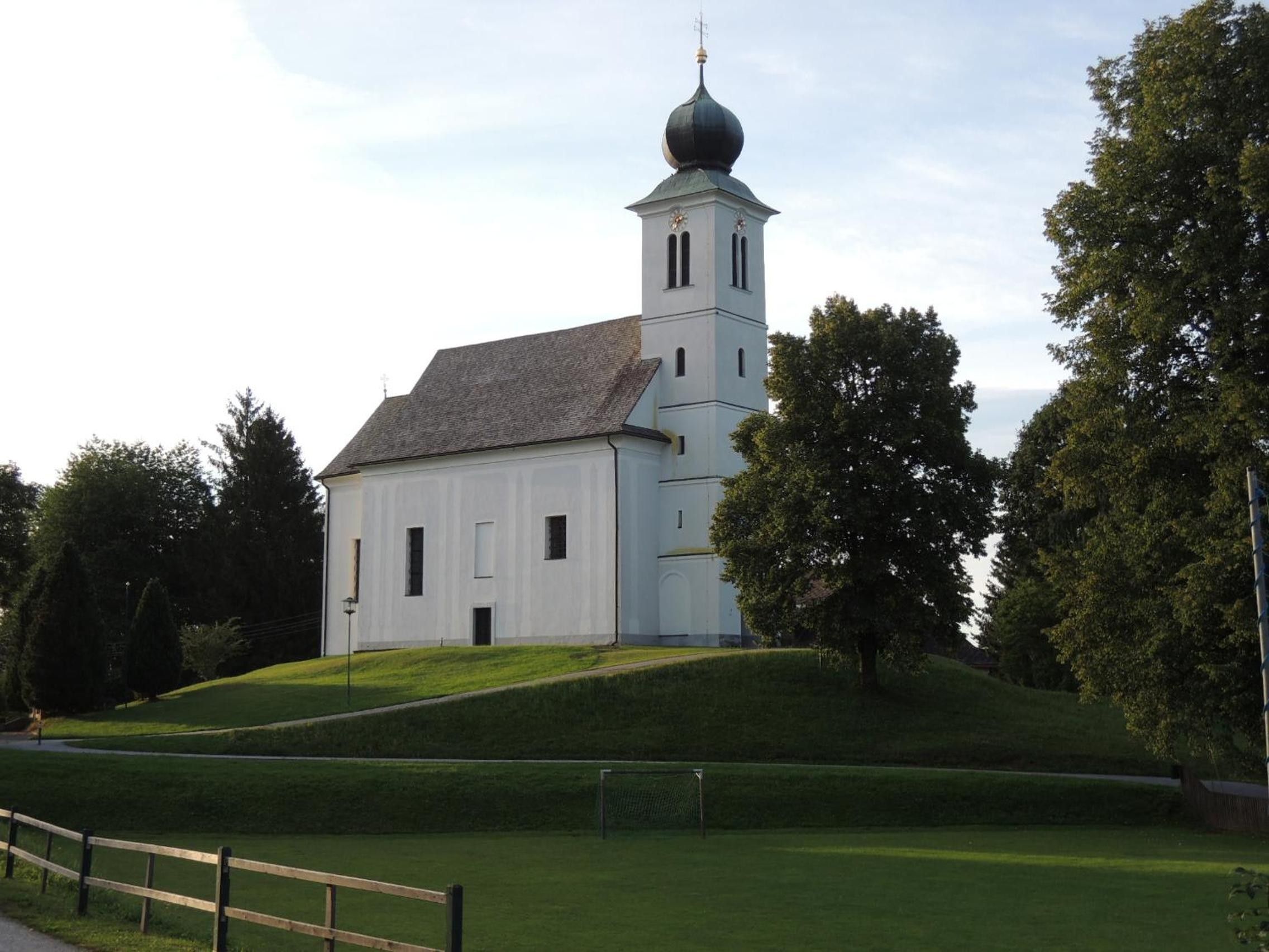 A white church with a bell tower and a dome, surrounded by trees and a grassy hill.
