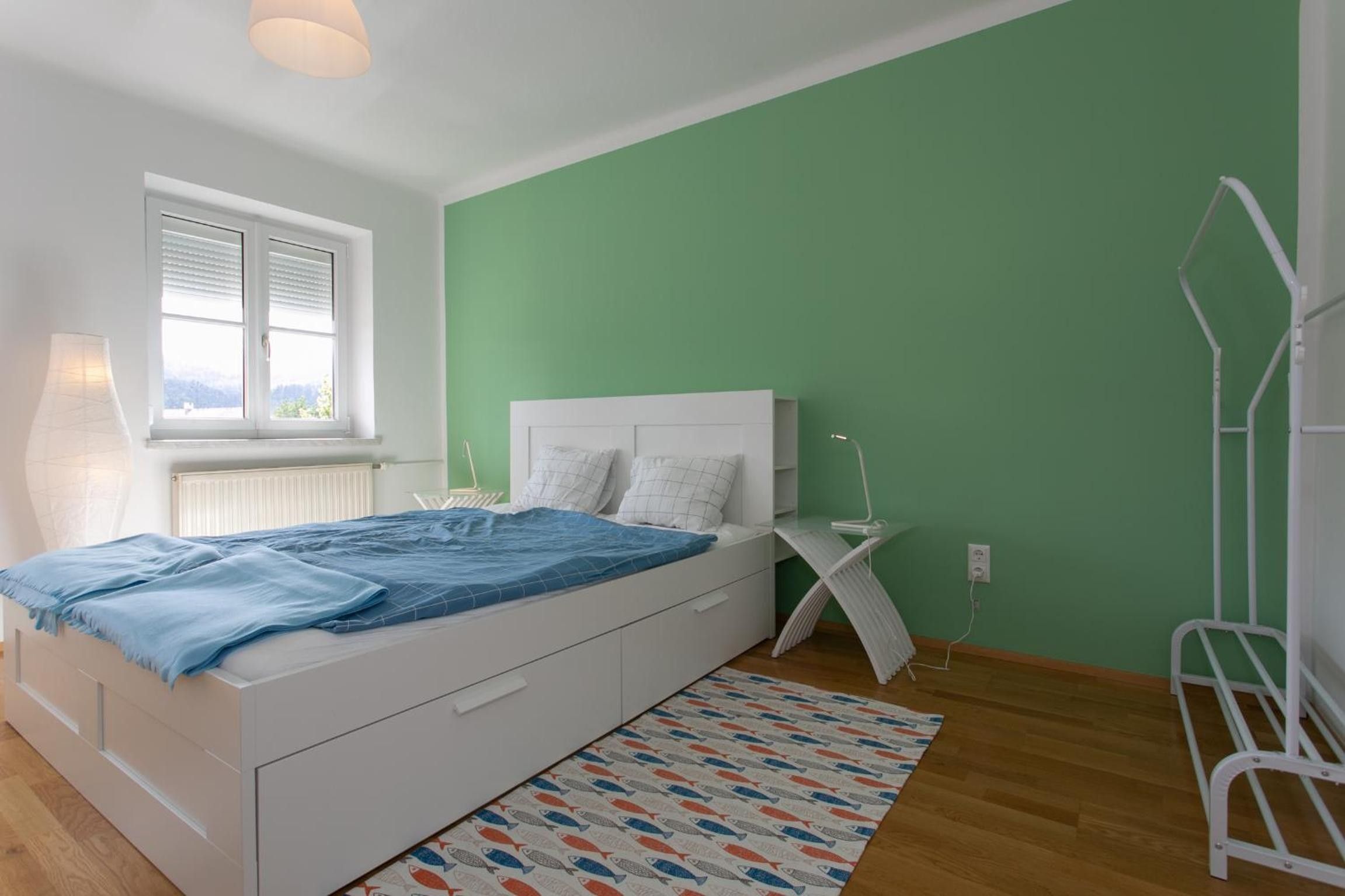 Un dormitorio con cama, ventana, radiador, mesita de noche y pared verde.