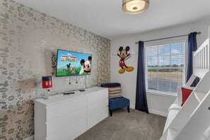 A child's room with a television displaying 'Mickey Mouse Clubhouse', a Mickey Mouse wall decal, a white dresser, a blue bench, and a window with curtains. - Oasis Villa Retreat at Champions Gate (Davenport)