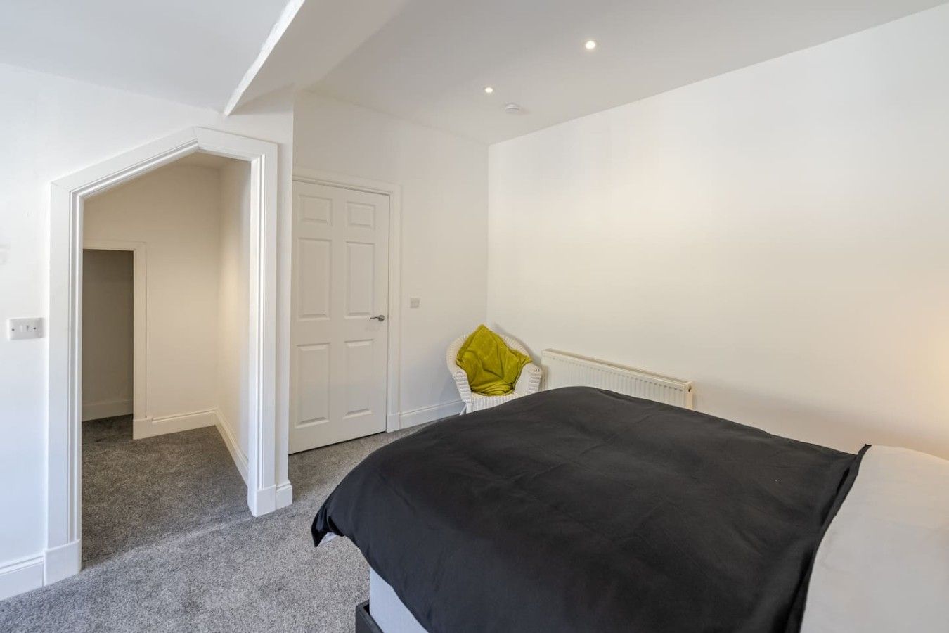 A bedroom with a bed, a white door, a radiator, and a chair with a yellow cushion.