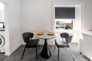 A small room with a round table, four chairs, a radiator, a window with a view of buildings, and a washing machine. - Newmarket Suites 21 in Ayr (South Ayrshire)
