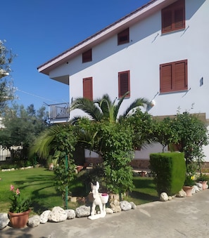 Ein weißes Gebäude mit roten Fensterläden, einem Balkon und einem gepflegten Garten mit Statue und Pflanzkübeln.