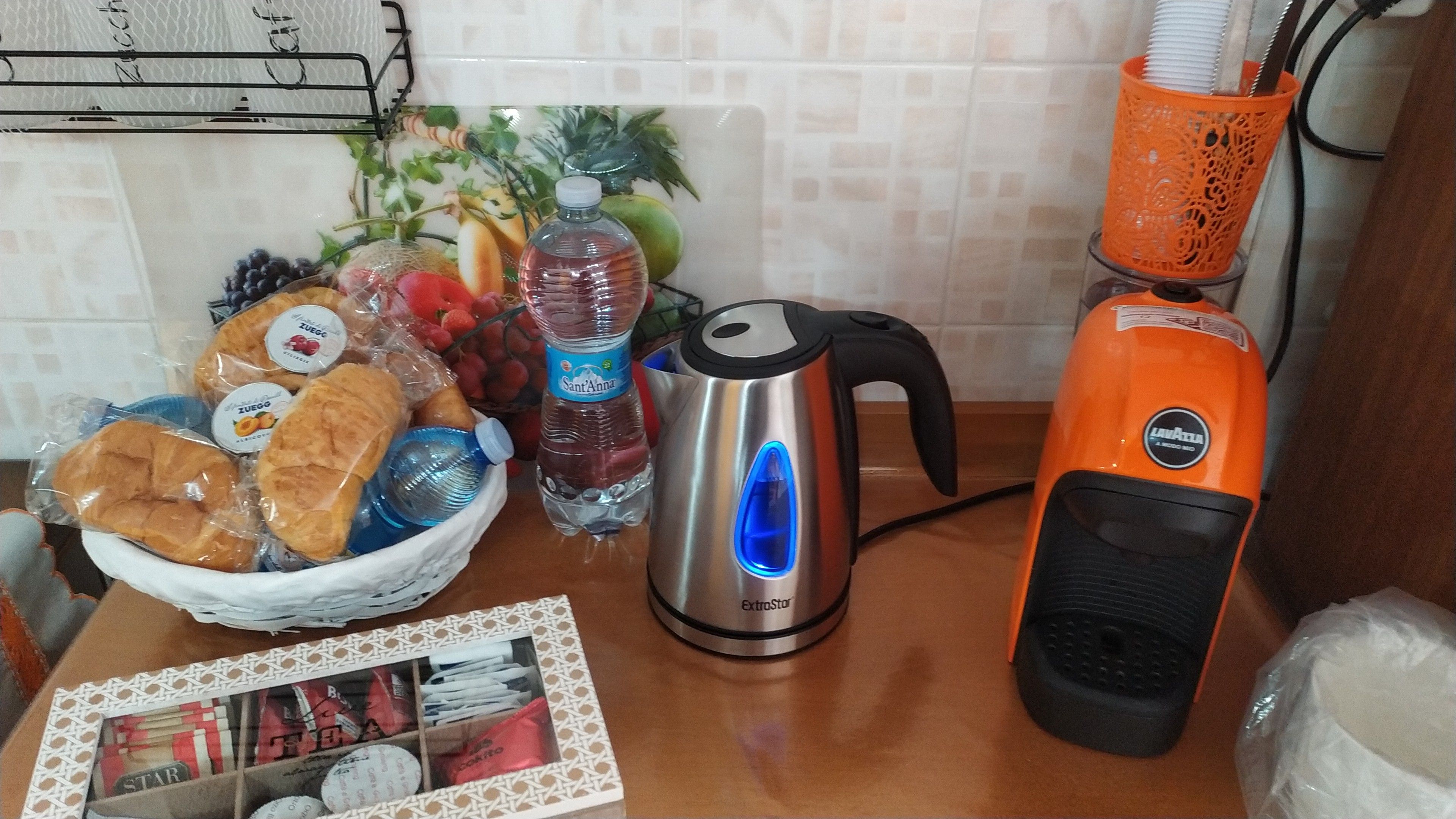 Una encimera de cocina con un hervidor, una botella de agua, una cesta de pan y un frutero.