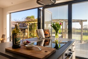Une cuisine moderne avec une grande fenêtre, une bouteille d’huile d’olive, une planche à découper et un vase rempli de fleurs.