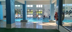 An indoor swimming pool with clear glass walls, blue pillars, and a lifebuoy on the right. - Modern Apartment with Ocean View Near Downtown in a Hotel Complex (Albufeira)