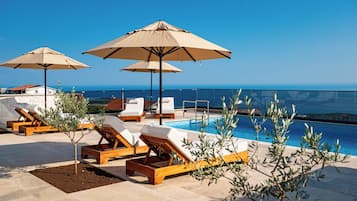 Een zwembadterras met ligbedden en parasols, met uitzicht op een helderblauwe lucht en een verre kustlijn.