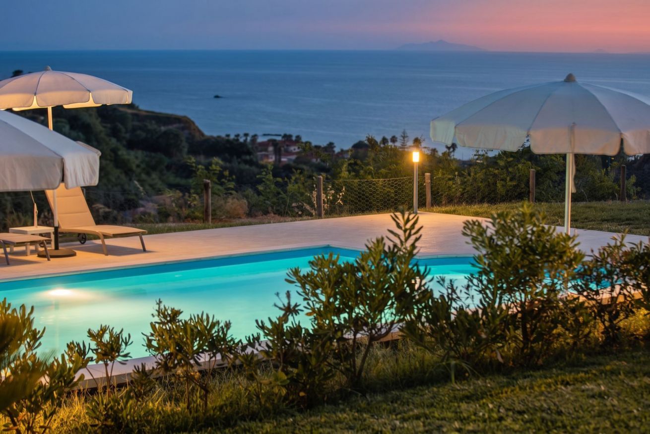 Ein Swimmingpool mit Liegestühlen und Sonnenschirmen, umgeben von Grünflächen mit Blick auf ein Gewässer.