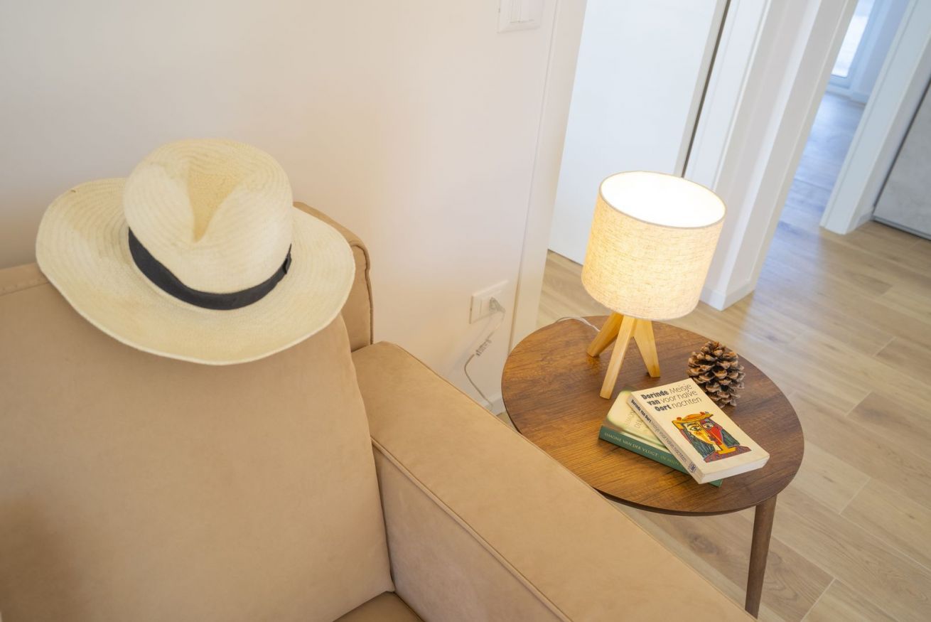 A room with a beige sofa, a small wooden side table with a lamp, and a book.