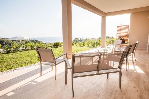 Un espace extérieur aménagé, abrité et doté de sièges, offrant une vue sur la mer et la verdure.