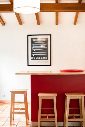 A kitchen area with a red accent wall, wooden stools, a framed picture of surfboards, and a red bowl on the counter. - Villa des Landes - familiale - Seignosse (Seignosse)