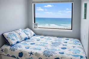 A bedroom with a view of the ocean, a bed with a blue and white ocean-themed bedspread, and a window with a view of the beach. - Direct Oceanfront! Charming condo in Myrtle Beach perfect for your getaway! (Myrtle Beach)