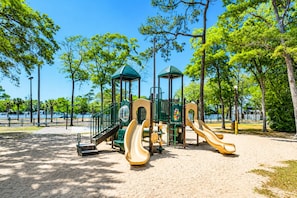 A playground with two slides and a climbing structure, surrounded by trees and a clear sky. - Direct Oceanfront! Charming condo in Myrtle Beach perfect for your getaway! (Myrtle Beach)