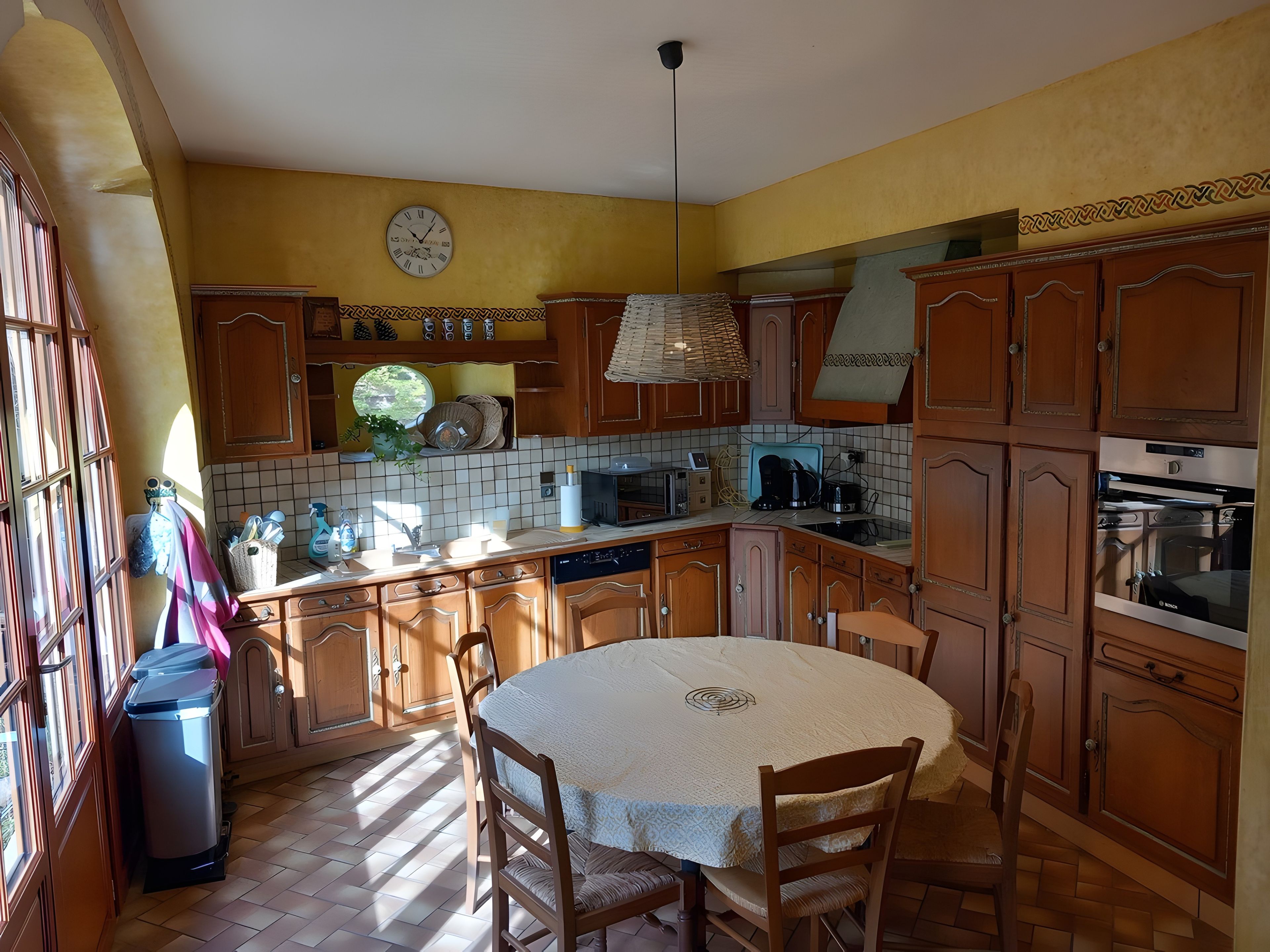 Een keuken met houten kastjes, een ronde eettafel en een klok aan de muur.