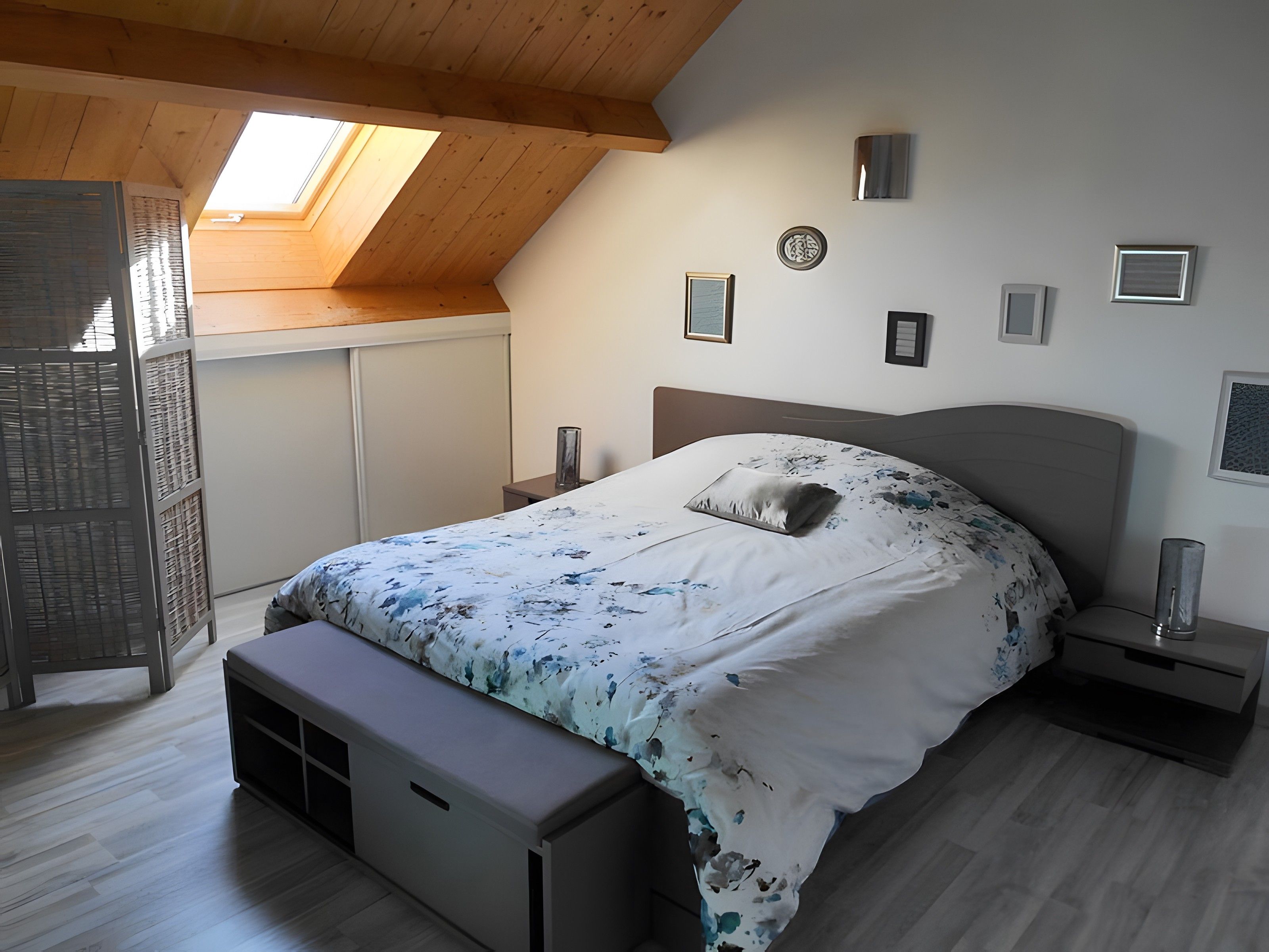 Un dormitorio con techo inclinado, tragaluces, cama con colcha floral, mesitas de noche y marcos de cuadros en la pared.