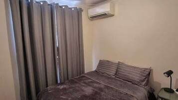 A bedroom with a bed, curtains, an air conditioning unit, and a nightstand with a lamp.