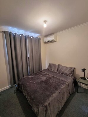 Een slaapkamer met een bed, gordijnen, een airconditioning en een nachtkastje met een lamp.