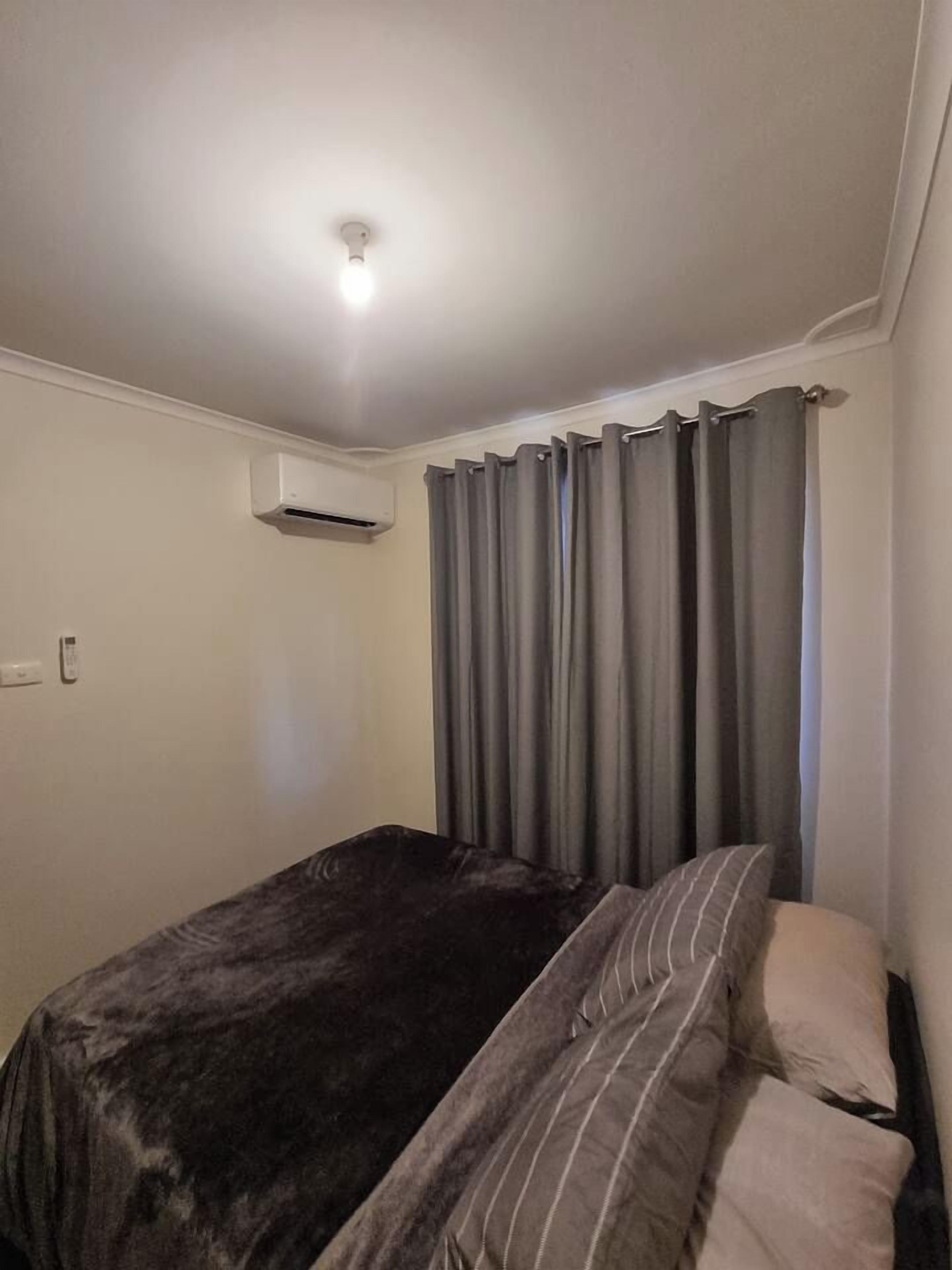 A small, dimly lit room with a single bed, a wall-mounted air conditioner, and grey curtains.
