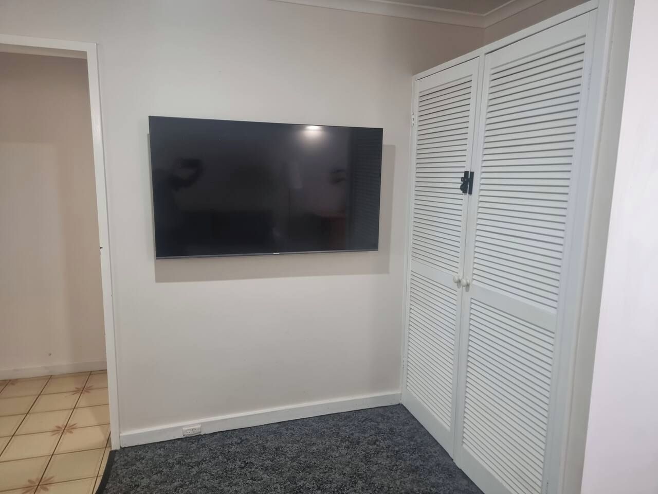 A room with a flat-screen TV mounted on the wall, a closed white closet with horizontal louvered doors, and a dark grey carpet.