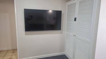 A room with a flat-screen TV mounted on the wall, a closed white closet with horizontal louvered doors, and a dark grey carpet.