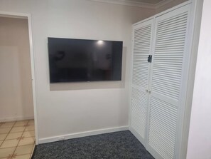Een kamer met een wandgemonteerde flatscreen televisie, een witte kast met horizontale verstelbare deuren en een donkergrijze vloerbedekking.
