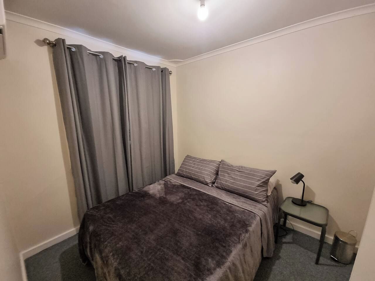 A bedroom with a bed, curtains, a bedside table with a lamp, and a trash bin.