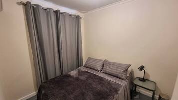 A bedroom with a bed, curtains, a bedside table with a lamp, and a trash bin.