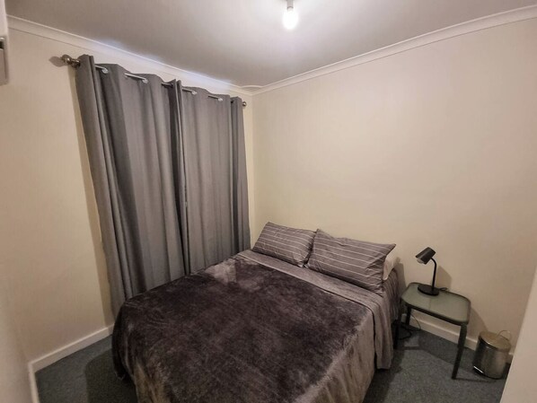 A bedroom with a bed, curtains, a bedside table with a lamp, and a trash bin.