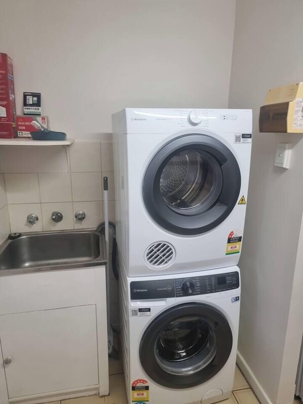 Un appareil combiné lave-linge et sèche-linge, de type « superposé », installé dans une petite buanderie équipée d’un évier et d’un meuble de rangement.