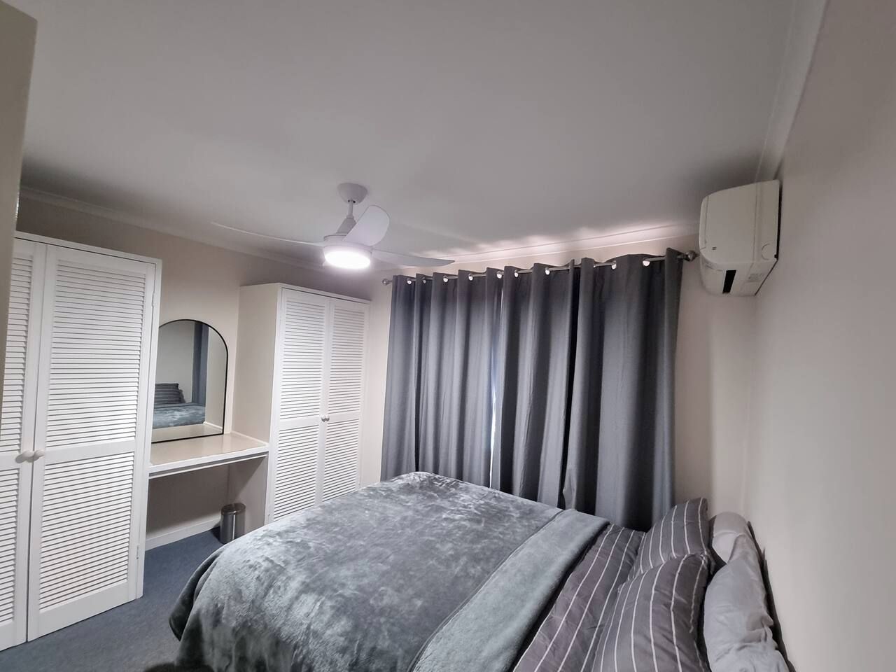 A bedroom with a bed, a bedside table, a mirror, a ceiling fan, and a window with curtains.