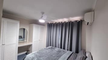 A bedroom with a bed, a bedside table, a mirror, a ceiling fan, and a window with curtains.