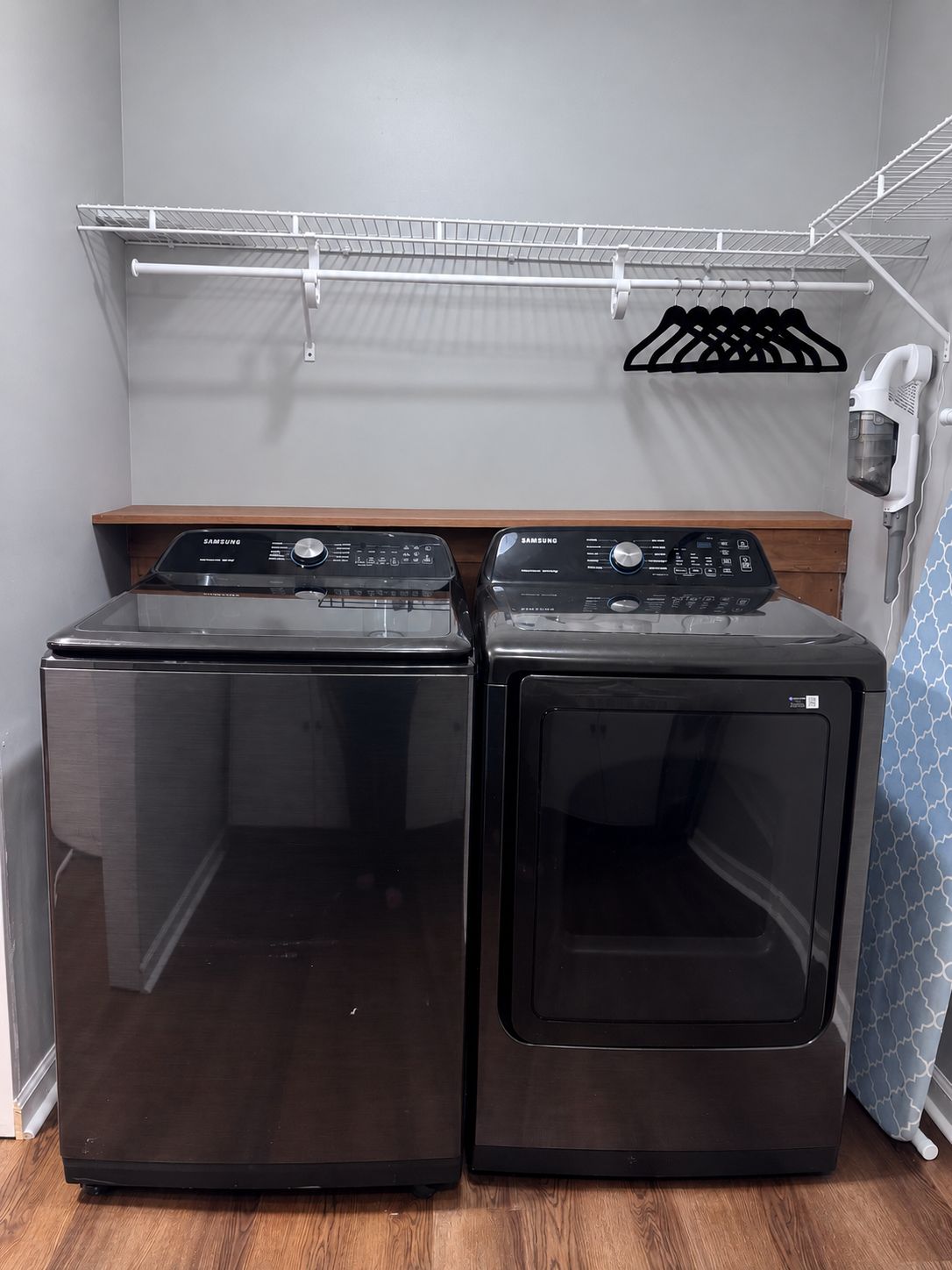 A laundry area with two front-loading washers and dryers, a metal shelf above, and a hanging rack with clothes.