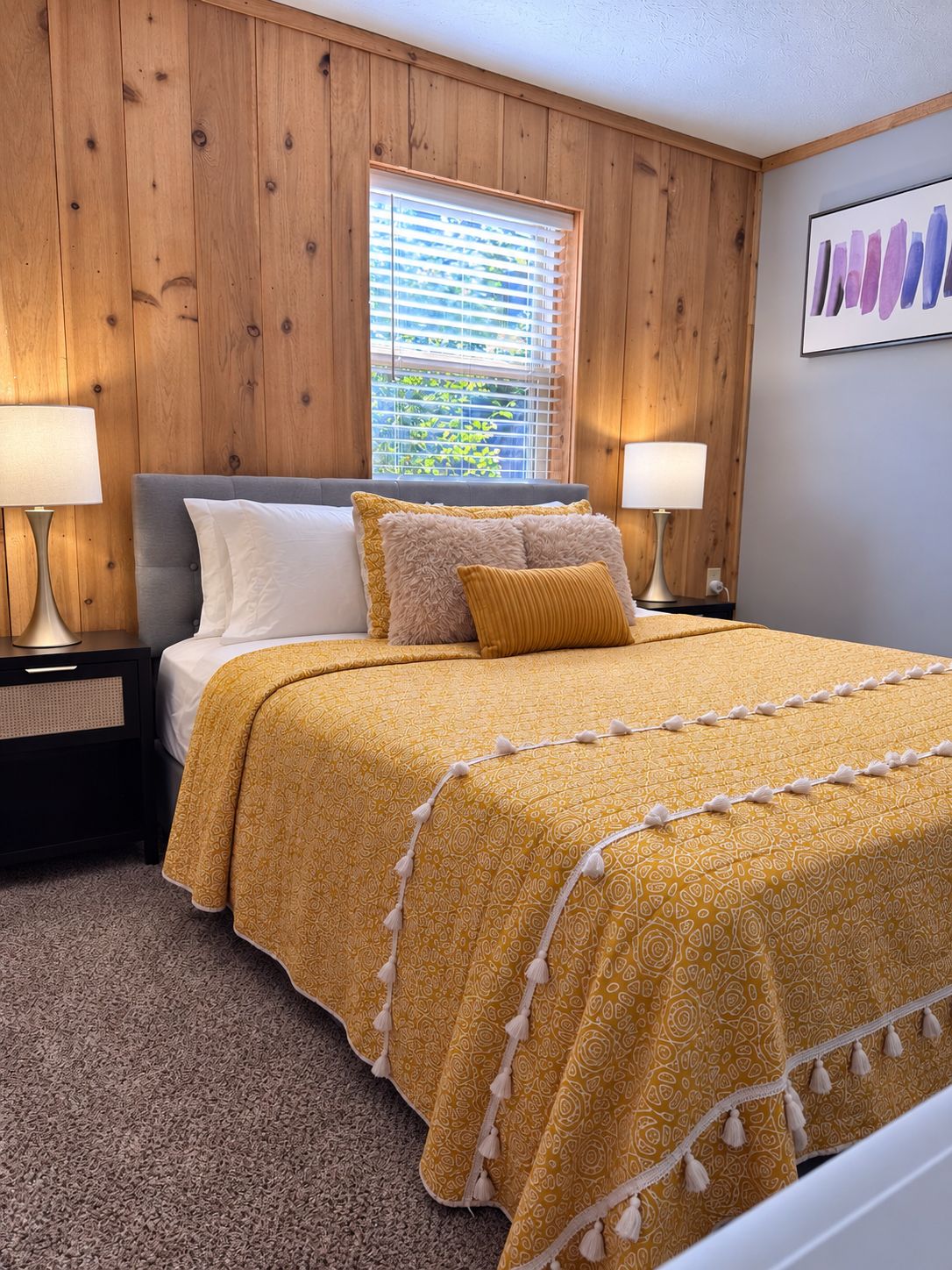 A bedroom with a bed, two bedside tables, a window with blinds, and a wall-mounted artwork.