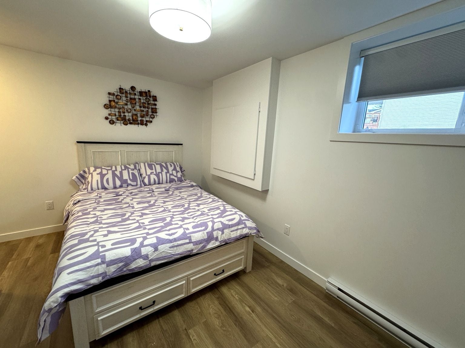 Ein Schlafzimmer mit einem Bett, einem an der Wand befestigten Regal, einem Fenster mit Jalousien und einer weißen Deckenleuchte.