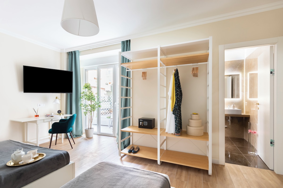 Ein modernes Schlafzimmer mit einem Flachbildfernseher, einem Schreibtisch mit Stuhl, einem Bett mit grauer Decke, einem Kleiderschrank mit Kleidung und einem Badezimmer mit Waschbecken und Spiegel.