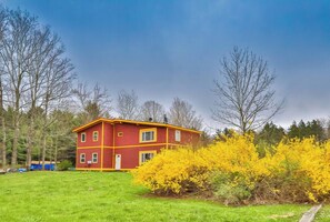 Een rode, twee verdiepingen tellende woning met een klein terras, omgeven door groen gras en bloeiende gele struiken.