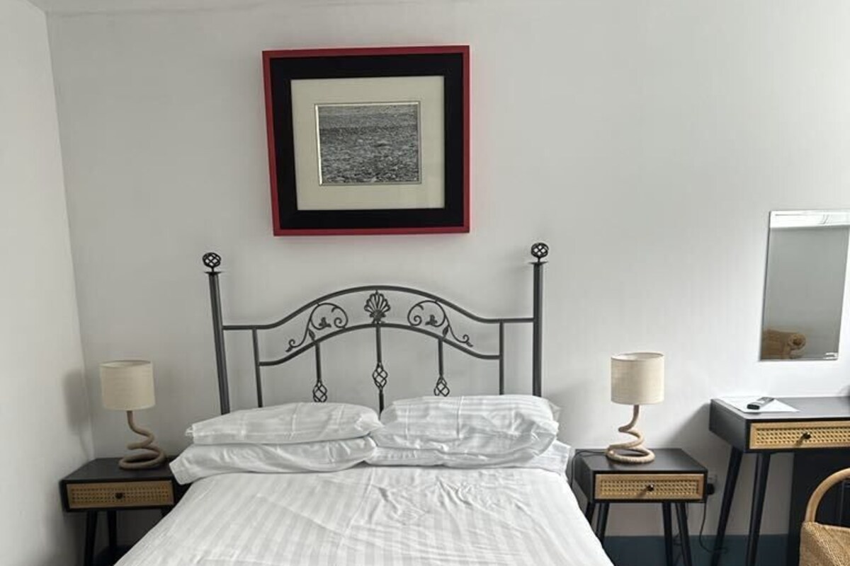 A neatly made bed with a headboard, two bedside tables with lamps, a desk with a chair, and a framed picture on the wall.