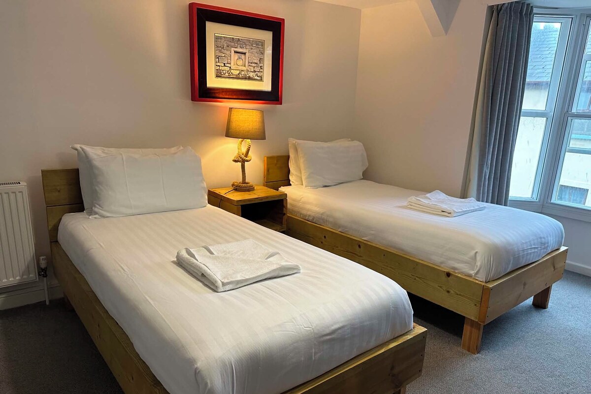 A hotel room with two wooden beds, white linens, a framed picture above the beds, a bedside lamp, and a window with curtains.