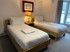 A hotel room with two wooden beds, white linens, a framed picture above the beds, a bedside lamp, and a window with curtains. - The Pier Hotel (Aberystwyth)